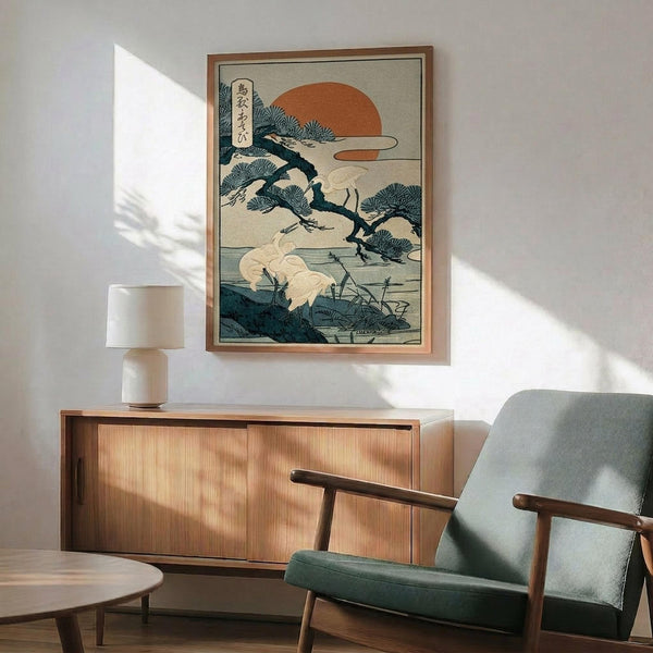 May include: Japanese woodblock print artwork featuring white cranes near water with pine trees and orange sun, displayed in wooden frame above mid-century modern walnut credenza. Green upholstered armchair with wooden arms sits nearby, white ceramic table lamp on credenza. Warm sunlight creates geometric shadows on white wall in minimalist living room setting.