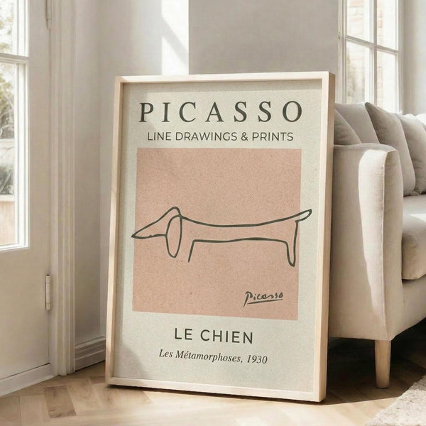 May include: Framed beige and peach poster featuring minimalist black line drawing of a dachshund dog in continuous line art style, displayed leaning against white wall next to light gray upholstered sofa in bright, airy room with large windows and natural lighting on wooden floors.