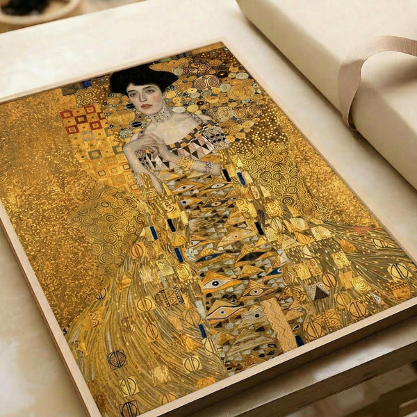 A reproduction print of Gustav Klimt's golden portrait painting showing a woman with dark hair wearing an ornate dress covered in geometric patterns and eye motifs. The artwork features Klimt's signature gold leaf style with intricate circular and rectangular designs filling the background. The print lies flat on white paper with rolls of paper visible in the background, displaying the rich golden tones and Byzantine-inspired decorative elements characteristic of Klimt's Golden Period work.
