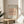May include: Framed Picasso-style minimalist line drawing of a dachshund dog on peachy-pink background with exhibition poster text, displayed on round wooden side table next to beige upholstered chair and plant in bright, airy room with white walls and natural sunlight shadows.