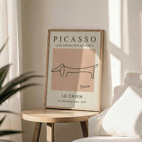 May include: Framed Picasso-style minimalist line drawing of a dachshund dog on peachy-pink background with exhibition poster text, displayed on round wooden side table next to beige upholstered chair and plant in bright, airy room with white walls and natural sunlight shadows.