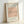 May include: Framed art print featuring Picasso's minimalist line drawing of a dachshund dog in black ink on a peachy-pink background. The poster has museum-style typography with "PICASSO LINE DRAWINGS & PRINTS" at top and "LE CHIEN Les Métamorphoses, 1930" at bottom. Displayed in light wooden frame leaning against white wall with soft natural lighting creating gentle shadows.