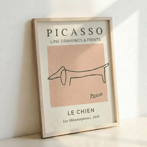 May include: Framed art print featuring Picasso's minimalist line drawing of a dachshund dog in black ink on a peachy-pink background. The poster has museum-style typography with "PICASSO LINE DRAWINGS & PRINTS" at top and "LE CHIEN Les Métamorphoses, 1930" at bottom. Displayed in light wooden frame leaning against white wall with soft natural lighting creating gentle shadows.