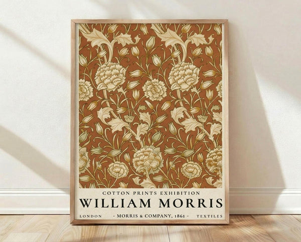 May include: Framed poster with William Morris floral pattern in warm brown and cream colors, featuring detailed botanical design with large blooming flowers and curving leaves. Light wooden frame rests against white textured wall on natural wood flooring with soft natural lighting creating gentle shadows.
