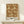 May include: Framed botanical poster with rust brown background featuring cream-colored floral pattern of large blooms, leaves and stems in William Morris style, displayed on light wood console table beside small white gift box with evergreen sprig against neutral wall