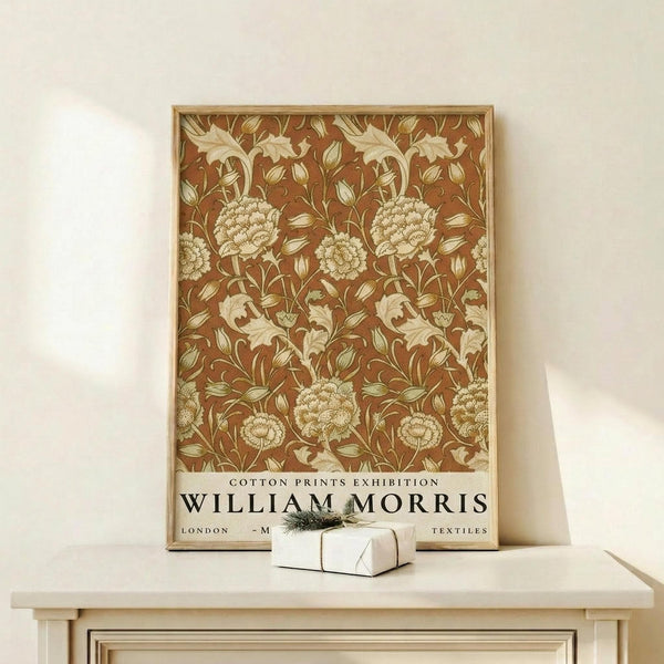 May include: Framed botanical poster with rust brown background featuring cream-colored floral pattern of large blooms, leaves and stems in William Morris style, displayed on light wood console table beside small white gift box with evergreen sprig against neutral wall