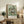 May include: A framed Van Gogh poster print featuring a lush bouquet of white roses in an earthenware vase against a mint green background with diagonal brushstrokes. The print has museum-style text below the artwork identifying the piece. The frame sits on a rustic wooden sideboard next to decorative pottery, ferns, books, and a beige upholstered chair with leather pillows in a warm, neutral-toned living space.