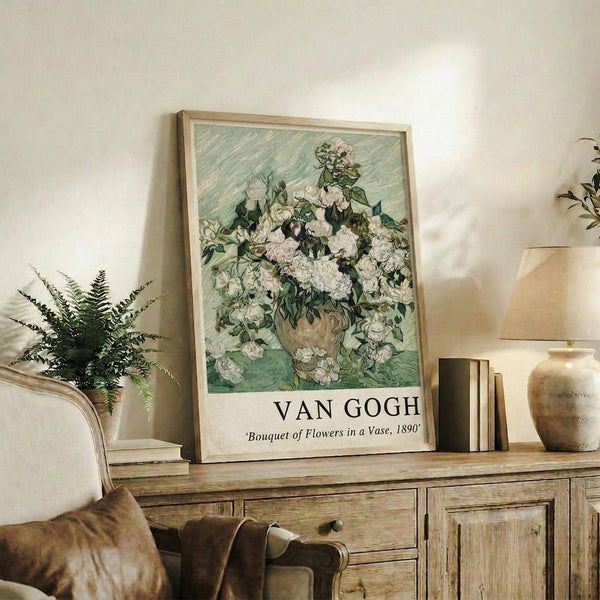 May include: A framed Van Gogh poster print featuring a lush bouquet of white roses in an earthenware vase against a mint green background with diagonal brushstrokes. The print has museum-style text below the artwork identifying the piece. The frame sits on a rustic wooden sideboard next to decorative pottery, ferns, books, and a beige upholstered chair with leather pillows in a warm, neutral-toned living space.