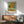 May include: Van Gogh's Sunflowers reproduction poster in thin brown frame hanging on white wall above wooden sideboard. Vibrant yellow sunflowers in vase against turquoise background with artist name and date below. Mid-century modern room with teal upholstered chair, round wood coffee table, white ceramic lamp. Natural sunlight creates diagonal shadows across wall and furniture.