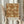 May include: Person holding framed vintage botanical print featuring ornate rust-colored background with cream and gold floral pattern of large blooms, leaves, and curving vines. Light wood frame against neutral curtained background with dried pampas grass visible to left side.