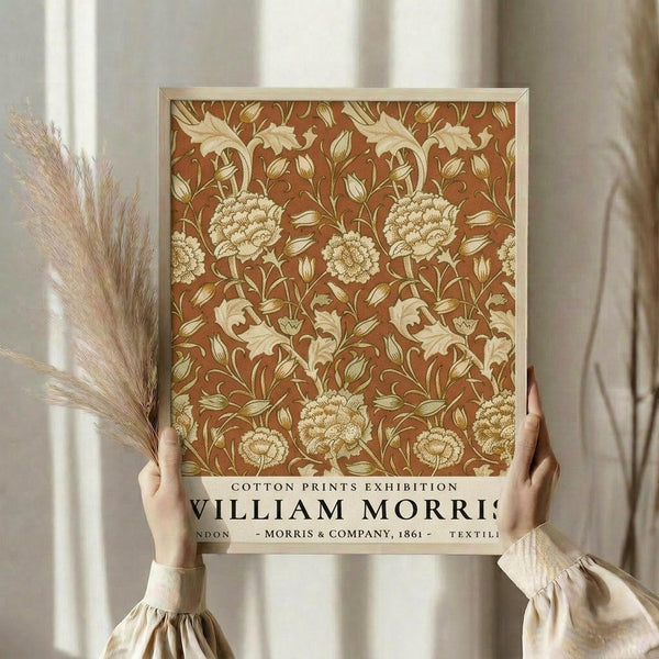 May include: Person holding framed vintage botanical print featuring ornate rust-colored background with cream and gold floral pattern of large blooms, leaves, and curving vines. Light wood frame against neutral curtained background with dried pampas grass visible to left side.
