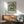 May include: Van Gogh poster print of white roses in brown vase against green brushstroke background, displayed in wooden frame above mid-century modern credenza. Scene shows mint green upholstered armchair with wooden arms, white ceramic lamp, and round coffee table. Warm natural light creates geometric shadows on white wall in minimalist living room setting.