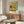 May include: Van Gogh's Sunflowers poster print in black frame hanging above wooden sideboard in modern living room. Yellow sunflowers in vase against turquoise background with white text below. Round white chair and glass coffee table visible. Natural lighting creates warm shadows on neutral walls.