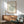 May include: Framed art print featuring black line drawing of leafy bush or shrub on cream background with "MATISSE GALERIE MAEGHT" text, displayed above wooden sideboard with ceramic table lamp, next to sage green mid-century armchair with wooden arms in sunlit room with white walls