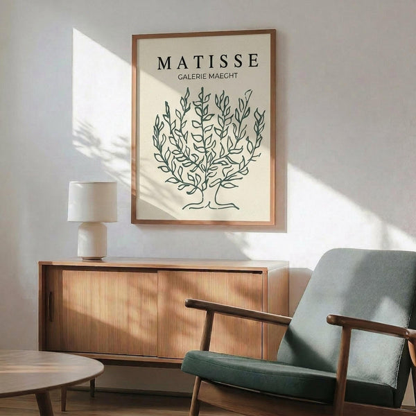 May include: Framed art print featuring black line drawing of leafy bush or shrub on cream background with "MATISSE GALERIE MAEGHT" text, displayed above wooden sideboard with ceramic table lamp, next to sage green mid-century armchair with wooden arms in sunlit room with white walls