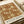 Vintage-style poster featuring William Morris floral textile pattern in warm rust brown with cream and gold botanical motifs including detailed flowers, leaves, and vines. The decorative design covers most of the print with "WILLIAM MORRIS" text and "COTTON PRINTS EXHIBITION" along with "LONDON - MORRIS & COMPANY, 1861 - TEXTILES" at bottom. Poster sits on cream fabric surface with rolled papers and beige ribbon visible nearby.