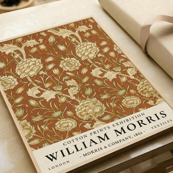 Vintage-style poster featuring William Morris floral textile pattern in warm rust brown with cream and gold botanical motifs including detailed flowers, leaves, and vines. The decorative design covers most of the print with "WILLIAM MORRIS" text and "COTTON PRINTS EXHIBITION" along with "LONDON - MORRIS & COMPANY, 1861 - TEXTILES" at bottom. Poster sits on cream fabric surface with rolled papers and beige ribbon visible nearby.