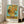 May include: Framed poster print of Van Gogh's famous sunflowers painting displayed in bright living room. Shows vibrant yellow sunflowers in ceramic vase against turquoise background, with artist name and date below artwork. Light wood frame leans against white wall next to beige sofa and large windows with natural sunlight streaming in.