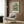 May include: Van Gogh floral poster print in light wood frame mounted on warm brown wood paneled wall above cream upholstered armchair with geometric patterned pillow. Golden brass floor lamp with white circular base stands nearby next to dark round side table with open book. Soft natural lighting from window with sheer curtains creates cozy modern reading nook atmosphere.