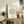 May include: Framed beige poster with black line drawing of leafy bush or tree, titled "MATISSE GALERIE MAEGHT" and "LE BUISSON Henri Matisse, 1951" in black text. Light wood frame sits on rustic wooden sideboard with drawers and cabinet doors. Scene includes beige armchair with brown leather pillow, potted fern, ceramic vases, stacked books, and trailing ivy plant hanging from above. Warm neutral tones throughout cozy living room setting.