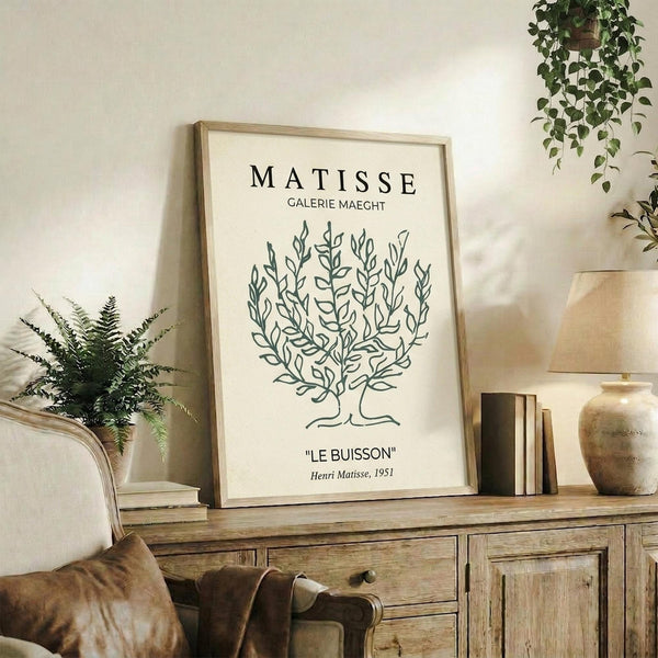 May include: Framed beige poster with black line drawing of leafy bush or tree, titled "MATISSE GALERIE MAEGHT" and "LE BUISSON Henri Matisse, 1951" in black text. Light wood frame sits on rustic wooden sideboard with drawers and cabinet doors. Scene includes beige armchair with brown leather pillow, potted fern, ceramic vases, stacked books, and trailing ivy plant hanging from above. Warm neutral tones throughout cozy living room setting.