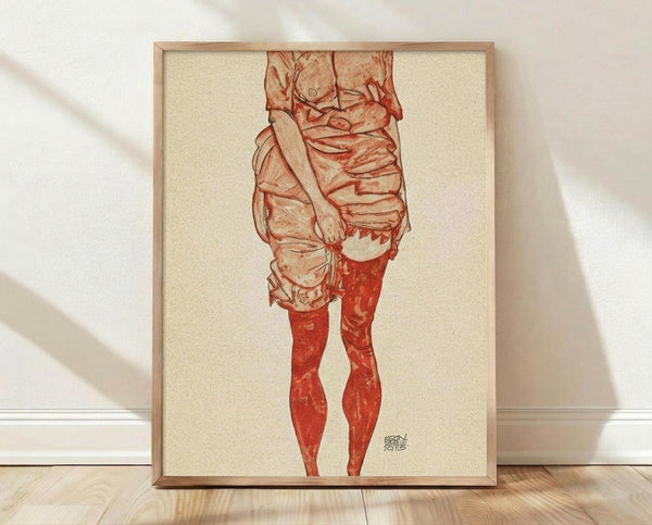 May include: Framed reproduction of an Egon Schiele drawing showing the lower torso and legs of a standing figure in red and sepia tones on cream paper, displayed in a light wood frame against white walls on wooden flooring with natural window light casting shadows.