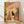 May include: Framed illustration print leaning against white wall on wooden floor. Artwork shows black cat sitting on toilet reading newspaper in colorful bathroom with pink tile walls, yellow curtain, green plants on shelf, small framed picture, and checkered floor. Warm lighting creates shadows on wall. Natural wood frame surrounds the whimsical domestic scene rendered in soft, muted tones.