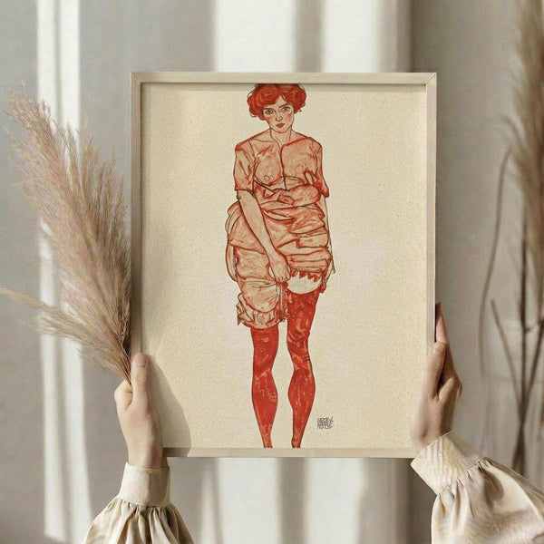 May include: Hands holding a framed art print featuring a figure with curly red hair wearing red stockings and draped fabric, drawn in an expressive linear style with reddish-brown tones on cream paper. The wooden frame is displayed against white curtains with dried wheat stalks visible, creating a neutral aesthetic room setting.
