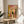 May include: Framed bathroom illustration showing black cat sitting on toilet reading newspaper, surrounded by coral pink tiled walls, potted plants on shelf, yellow shower curtain, and colorful checkered floor. Art print displayed on round wooden side table next to beige upholstered chair in bright sunlit room with white walls and plant shadows.
