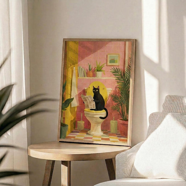 May include: Framed bathroom illustration showing black cat sitting on toilet reading newspaper, surrounded by coral pink tiled walls, potted plants on shelf, yellow shower curtain, and colorful checkered floor. Art print displayed on round wooden side table next to beige upholstered chair in bright sunlit room with white walls and plant shadows.