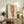 May include: Framed artwork displaying a figure study in red and brown tones, shown in a warm living room setting on a rustic wooden sideboard alongside books, ceramic vases, and a fern plant, with a beige upholstered chair visible in the foreground.