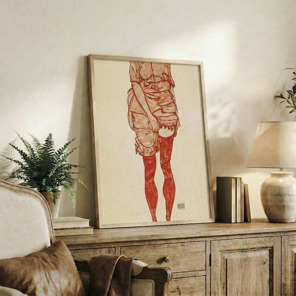 May include: Framed artwork displaying a figure study in red and brown tones, shown in a warm living room setting on a rustic wooden sideboard alongside books, ceramic vases, and a fern plant, with a beige upholstered chair visible in the foreground.