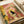 May include: Vintage-style illustration print displayed in open book or portfolio showing black cat sitting on yellow toilet reading newspaper in retro pink-tiled bathroom with houseplants, framed artwork on walls, and warm coral and yellow color palette in mid-century modern artistic style