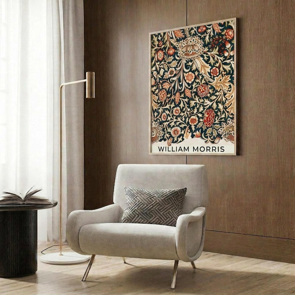May include: William Morris botanical poster with intricate floral pattern in navy blue background featuring ornate flowers, leaves, and vines in coral, cream, and gold tones. Displayed in light wood frame on textured wood panel wall above modern beige upholstered armchair with geometric patterned pillow. Room includes brass floor lamp and dark side table with open book.