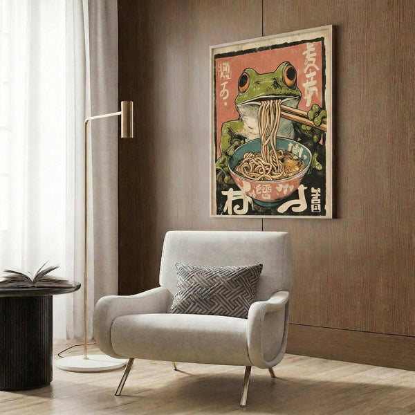 May include: Japanese-style vintage poster featuring a green frog eating ramen noodles from a teal bowl, displayed in a light wooden frame against wood paneling. The artwork shows coral pink background with Japanese text characters. Scene includes modern beige armchair with geometric pillow, brass floor lamp, and dark round side table with open book.
