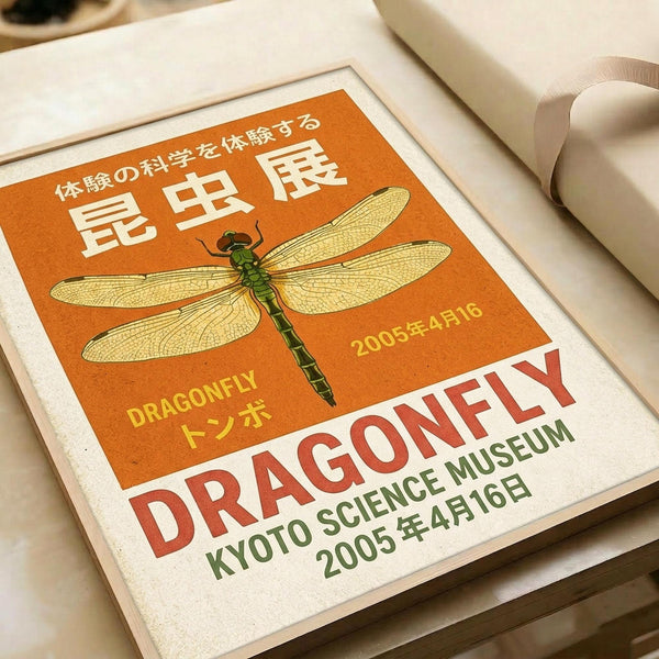 May include: Vintage-style poster print with orange background featuring detailed green dragonfly illustration and Japanese text. Shows "DRAGONFLY" and "KYOTO SCIENCE MUSEUM 2005年4月16日" in red and green lettering. Print displayed in white matted frame on light wooden surface next to rolled paper tubes, creating warm natural lighting and artistic flat lay composition.