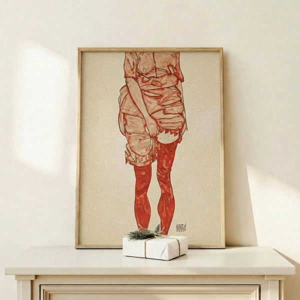 May include: Framed artwork displaying a figure study in red and orange tones, showing a person from torso to feet wearing draped clothing and red stockings, rendered in an expressive sketch style. The piece sits on a light wood surface beside a small wrapped gift with greenery, against a cream-colored wall in soft natural lighting.