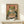 May include: A framed vintage-style Japanese poster featuring a green frog with orange eyes eating noodles from a colorful bowl using chopsticks. The artwork has a retro aesthetic with Japanese text on a coral and black background. The wooden-framed print sits on a light wood shelf next to a small white geometric object against a cream wall.