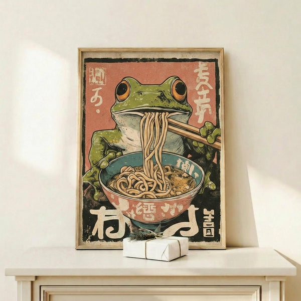 May include: A framed vintage-style Japanese poster featuring a green frog with orange eyes eating noodles from a colorful bowl using chopsticks. The artwork has a retro aesthetic with Japanese text on a coral and black background. The wooden-framed print sits on a light wood shelf next to a small white geometric object against a cream wall.