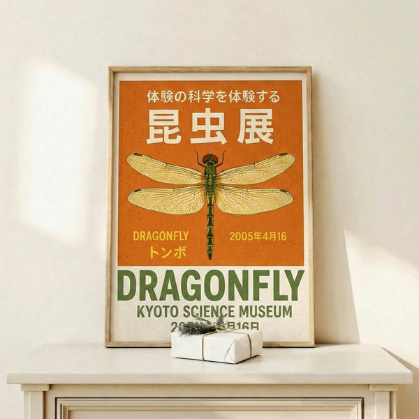 May include: Framed poster with orange and green design featuring detailed dragonfly illustration with translucent wings displayed on light wooden shelf against white wall. Japanese text at top, "DRAGONFLY" and "KYOTO SCIENCE MUSEUM" with date 2005年4月16日 below. Small wrapped gift box with evergreen sprig sits in front of natural wood frame.