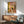 May include: Framed poster print featuring a black cat with yellow halo sitting on white toilet reading newspaper in colorful bathroom with pink tiled walls, plants on shelves, and checkered floor. Shown hanging above wood credenza with white table lamp in mid-century modern living room with sage green armchair and round coffee table.
