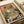 Vintage-style Japanese poster print featuring a green frog with large orange eyes eating noodles from a ramen bowl using chopsticks. The illustration has a retro color palette with coral pink, teal, and cream tones, surrounded by Japanese text characters. The artwork is displayed on white paper with a subtle textured appearance, positioned on what appears to be a light wooden surface.