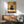 May include: Framed artwork showing a large black cat with bright yellow eyes sitting at a white table with pizza and teapot, displayed above a wooden sideboard with white lamp in a mid-century modern living room with sage green armchair and natural lighting creating shadows on white walls.