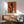 May include: Framed art print on white wall showing orange background with black silhouette of elegant woman holding wine glass and smaller light-colored figure also with glass. Wood-framed poster hangs above mid-century modern walnut credenza with white ceramic lamp. Sage green upholstered chair with wooden arms sits nearby on hardwood floor with natural sunlight creating dramatic shadows across the minimalist living room scene.