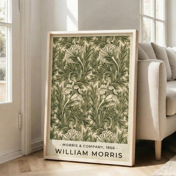 May include: Framed botanical art print featuring intricate green and cream acanthus leaf pattern with flowing, ornate foliage design. The vintage-style poster displays "Morris & Company, 1866" and "William Morris" text at bottom. Light wood frame leans against white wall beside beige upholstered chair in bright, minimalist living room with large windows and natural lighting.