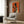 May include: Art deco style poster with orange background showing black silhouette of woman holding wine glass alongside smaller illustrated woman with blonde hair also holding glass, displayed in light wood frame on wood paneled wall above cream upholstered armchair with geometric pillow, brass floor lamp and side table visible in modern living room setting.