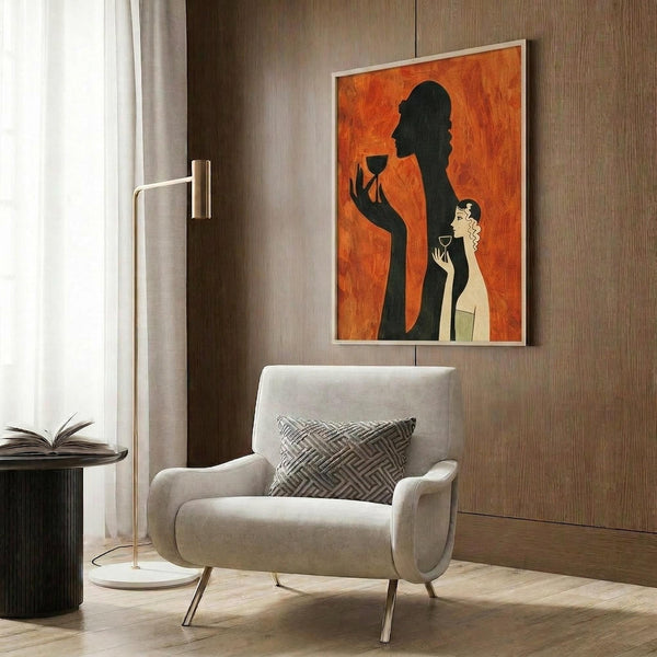 May include: Art deco style poster with orange background showing black silhouette of woman holding wine glass alongside smaller illustrated woman with blonde hair also holding glass, displayed in light wood frame on wood paneled wall above cream upholstered armchair with geometric pillow, brass floor lamp and side table visible in modern living room setting.