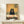 May include: Framed artwork displayed on white wooden console table showing black cat with wide yellow eyes eating pizza slice at checkered table in warm kitchen setting with yellow walls and framed art. White gift box with evergreen sprig and white ceramic vase with dried palm fronds styled nearby.