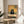 May include: Framed poster print displayed on round wooden side table next to beige upholstered chair and plant. Artwork shows black cat with wide yellow eyes sitting at checkered tablecloth-covered table with pizza slice, rendered in warm vintage illustration style with yellow wall background. Natural lighting creates soft shadows across white wall.