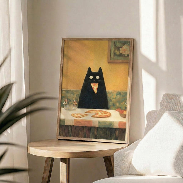 May include: Framed poster print displayed on round wooden side table next to beige upholstered chair and plant. Artwork shows black cat with wide yellow eyes sitting at checkered tablecloth-covered table with pizza slice, rendered in warm vintage illustration style with yellow wall background. Natural lighting creates soft shadows across white wall.